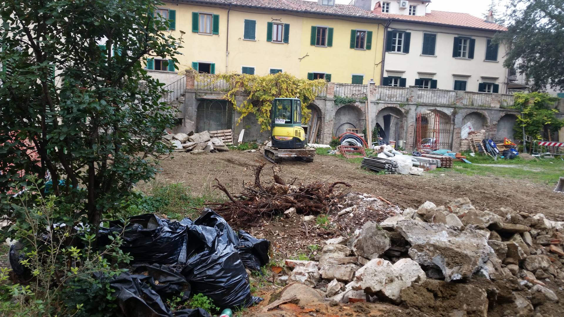 Cantiere esterno con escavatore giallo e cumuli di terra davanti al loggiato